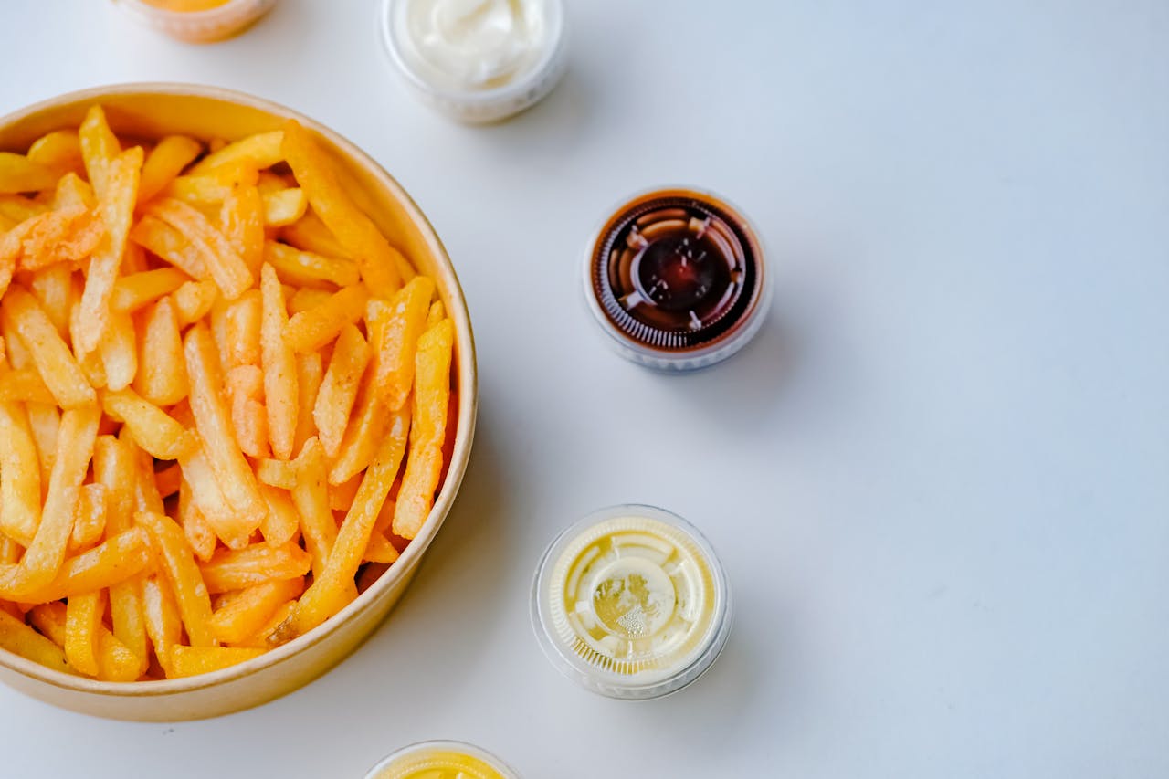 A top-view of a bowl of crispy French fries accompanied by various dipping sauces on a white surface.